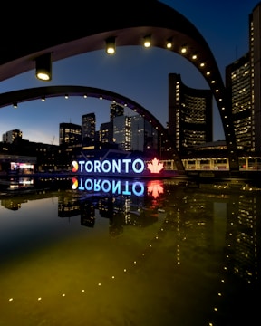 A night view of the Toronto city sign and skyline, featuring illuminated buildings reflected in a calm body of water. The large iconic 'TORONTO' sign is brightly lit, incorporating a red maple leaf symbol. The structures around are adorned with light archways, adding to the urban aesthetic.