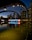 A night view of the Toronto city sign and skyline, featuring illuminated buildings reflected in a calm body of water. The large iconic 'TORONTO' sign is brightly lit, incorporating a red maple leaf symbol. The structures around are adorned with light archways, adding to the urban aesthetic.
