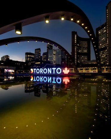 A night view of the Toronto city sign and skyline, featuring illuminated buildings reflected in a calm body of water. The large iconic 'TORONTO' sign is brightly lit, incorporating a red maple leaf symbol. The structures around are adorned with light archways, adding to the urban aesthetic.