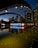 A night view of the Toronto city sign and skyline, featuring illuminated buildings reflected in a calm body of water. The large iconic 'TORONTO' sign is brightly lit, incorporating a red maple leaf symbol. The structures around are adorned with light archways, adding to the urban aesthetic.