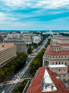 Aerial view of an urban area featuring roads lined with buildings. The architecture includes large governmental or institutional buildings with red-tiled roofs and rectangular designs. In the background, a prominent white dome structure stands out against the skyline. The sky is partly cloudy, with patches of blue visible.
