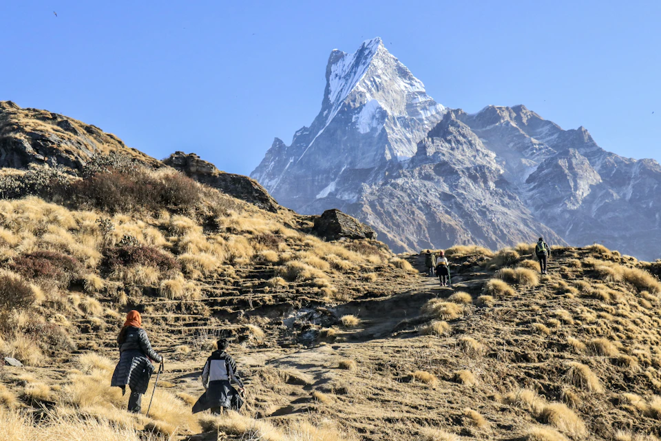 The image depicts a group of hikers ascending a mountain trail, with a majestic snow-capped peak visible in the background. The hikers are dressed in outdoor gear and carrying backpacks, suggesting they are well-prepared for their journey. The perspective is from ground level, looking up at the hikers as they make their way towards the top of the mountain.