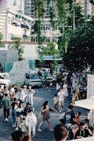 A lively outdoor scene with numerous people walking and mingling. Food trucks and vans are in the background, while tall residential and commercial buildings rise behind them. String lights hang above the people, adding to the festive atmosphere. Some individuals are wearing casual summer clothing, and a few children are present.