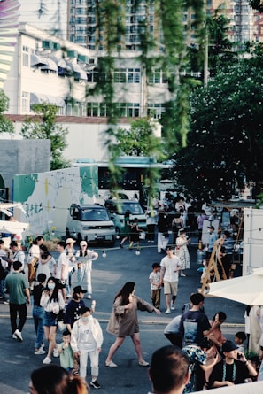 A lively outdoor scene with numerous people walking and mingling. Food trucks and vans are in the background, while tall residential and commercial buildings rise behind them. String lights hang above the people, adding to the festive atmosphere. Some individuals are wearing casual summer clothing, and a few children are present.