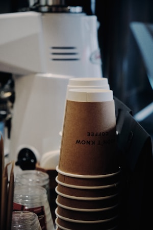 A stack of brown paper coffee cups neatly arranged, with a text partially visible on the top cup. In the background, a blurry white coffee machine is visible, alongside other coffee shop accessories such as glass jars and stir sticks.