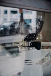 A close-up of a coffee grinder with visible coffee beans inside a transparent hopper. The grinder features a sleek white design with a metallic accent and logo label. The glass reflects part of the surrounding environment.