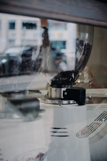 A close-up of a coffee grinder with visible coffee beans inside a transparent hopper. The grinder features a sleek white design with a metallic accent and logo label. The glass reflects part of the surrounding environment.