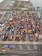 Aerial view of a bustling shipping container yard with rows of colorful containers stacked alongside orange cranes for loading and unloading. Roads with vehicles line the bottom of the image, and there's a body of water and open land in the background.