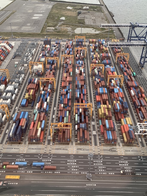 Aerial view of a bustling shipping container yard with rows of colorful containers stacked alongside orange cranes for loading and unloading. Roads with vehicles line the bottom of the image, and there's a body of water and open land in the background.