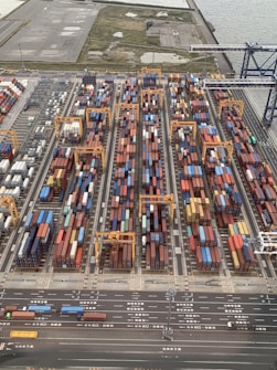 Aerial view of a bustling shipping container yard with rows of colorful containers stacked alongside orange cranes for loading and unloading. Roads with vehicles line the bottom of the image, and there's a body of water and open land in the background.