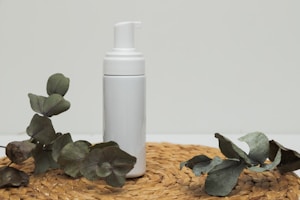 A white foam pump bottle is positioned upright on a woven straw mat. Surrounding the bottle are several green eucalyptus leaves, providing a natural and minimalist aesthetic.