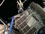 An astronaut in a white spacesuit floats outside a space structure, which appears intricate with panels and tubes. The background is a dark, starry space setting, emphasizing the feeling of being in outer space. The structure includes solar panels and metal trusses.