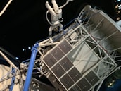 An astronaut in a white spacesuit floats outside a space structure, which appears intricate with panels and tubes. The background is a dark, starry space setting, emphasizing the feeling of being in outer space. The structure includes solar panels and metal trusses.