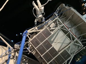 An astronaut in a white spacesuit floats outside a space structure, which appears intricate with panels and tubes. The background is a dark, starry space setting, emphasizing the feeling of being in outer space. The structure includes solar panels and metal trusses.