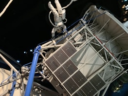 An astronaut in a white spacesuit floats outside a space structure, which appears intricate with panels and tubes. The background is a dark, starry space setting, emphasizing the feeling of being in outer space. The structure includes solar panels and metal trusses.
