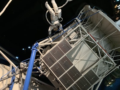 An astronaut in a white spacesuit floats outside a space structure, which appears intricate with panels and tubes. The background is a dark, starry space setting, emphasizing the feeling of being in outer space. The structure includes solar panels and metal trusses.