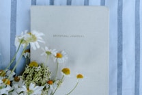 A spiritual journal with a beige cover lies on a fabric with blue and white stripes. Several white and yellow daisies are artfully arranged along the left side of the journal, adding a natural and calming touch.