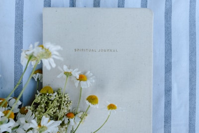 A spiritual journal with a beige cover lies on a fabric with blue and white stripes. Several white and yellow daisies are artfully arranged along the left side of the journal, adding a natural and calming touch.