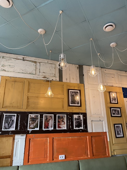 A creatively decorated wall features various colored panels and hanging lightbulbs with wireframes. The wall displays black and white photographs in different frames. The overall setting combines rustic textures with modern elements.