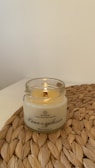 A lit candle is placed in a clear glass jar on a woven mat. The candle has a creamy color, and Russian text is printed on the jar. The background is a plain off-white wall.