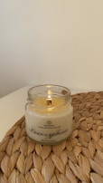 A lit candle is placed in a clear glass jar on a woven mat. The candle has a creamy color, and Russian text is printed on the jar. The background is a plain off-white wall.