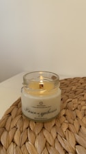 A lit candle is placed in a clear glass jar on a woven mat. The candle has a creamy color, and Russian text is printed on the jar. The background is a plain off-white wall.