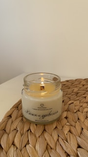 A lit candle is placed in a clear glass jar on a woven mat. The candle has a creamy color, and Russian text is printed on the jar. The background is a plain off-white wall.