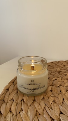 A lit candle is placed in a clear glass jar on a woven mat. The candle has a creamy color, and Russian text is printed on the jar. The background is a plain off-white wall.