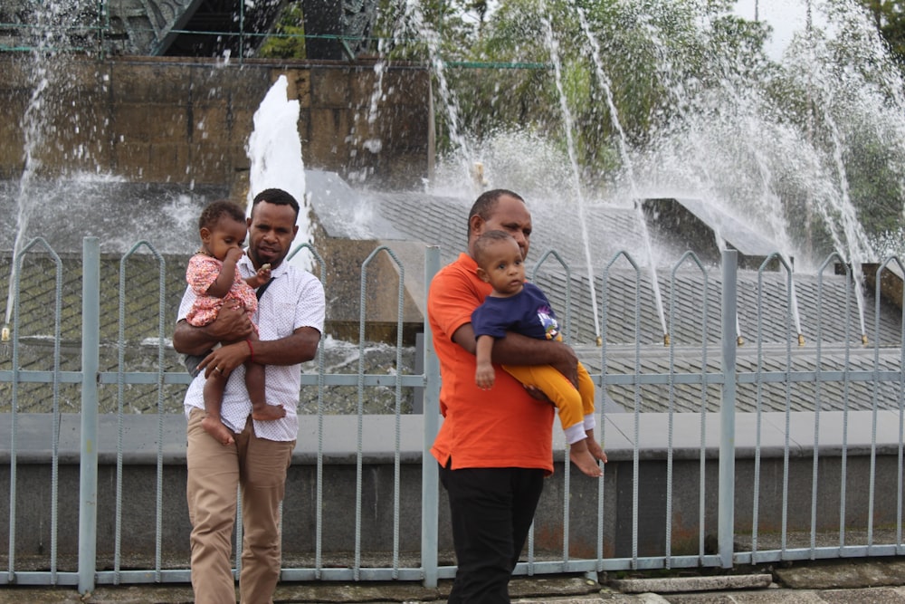 a person holding a baby and standing in front of a fountain