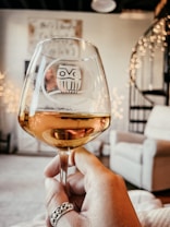 A hand holding a glass of amber-colored liquid, possibly wine, with a decorative owl design etched on the glass. In the background, there is a cozy interior setting with a white armchair and a spiral staircase. Warm fairy lights add a soft glow to the room.