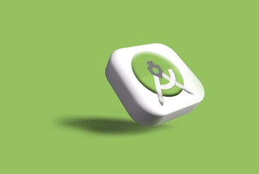 A three-dimensional icon with a green circular background and a white border, featuring a white compass or drafting tool at the center. The icon casts a slight shadow against a light green background.