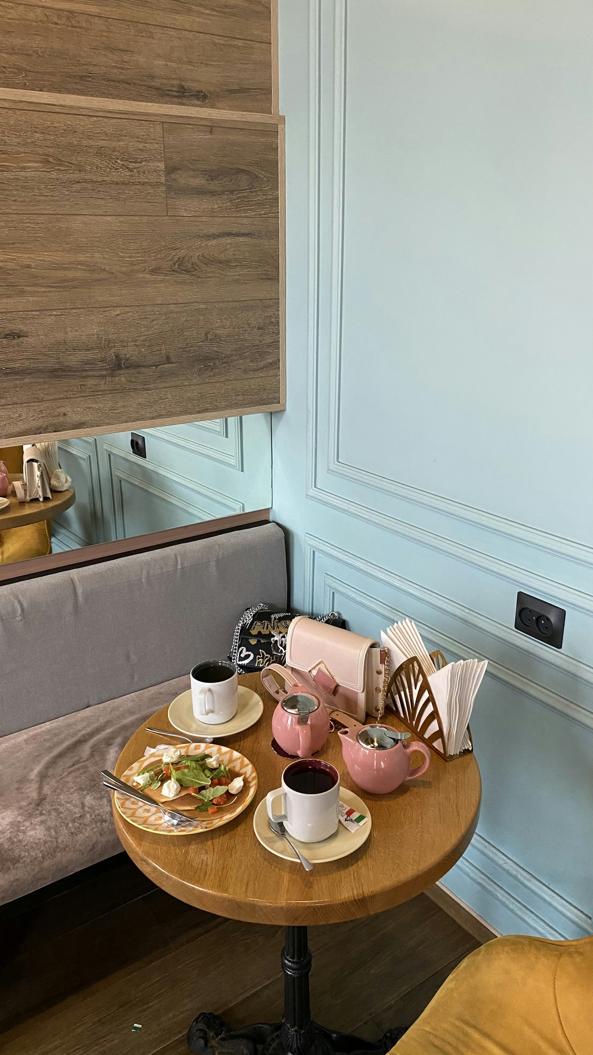 A cozy café setting with a small round wooden table. On the table are two pink teapots, two white ceramic cups filled with dark liquid, a plate of salad with lettuce and tomatoes, and a folded beige purse. The table is adjacent to a comfortable gray upholstered bench and situated against a light blue, paneled wall. Napkins are neatly arranged in a holder.