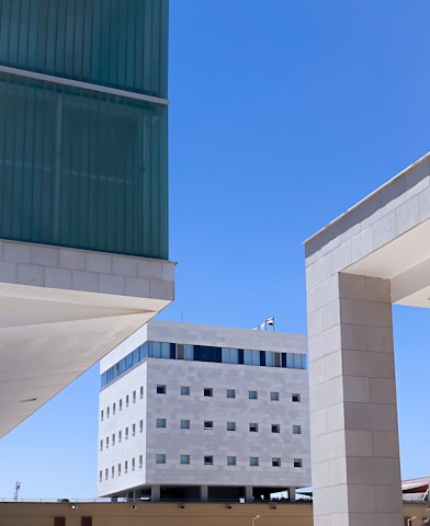 A modern architectural scene with multiple buildings featuring clean lines and geometric shapes. The structures have a mix of glass and stone materials, with the sky in a clear blue background. A distinct white building is centered in the background with a pattern of small square windows and a row of larger windows near the top.