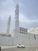 A large, white mosque with towering minarets and domed roofs. An overcast sky with a plane in the background. The mosque's architecture is predominantly white with intricate details. A white car is parked in the foreground, next to some street poles.