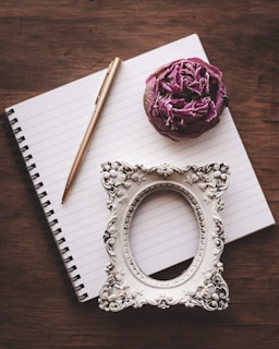 A spiral-bound notebook lies open on a wooden surface, accompanied by a gold pen, a dried purple rose, and an ornate empty picture frame with intricate designs.