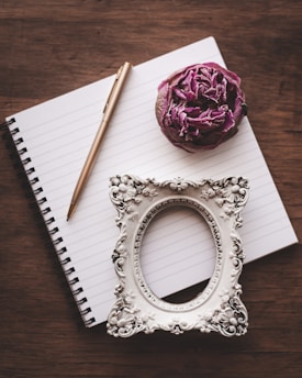 A spiral-bound notebook lies open on a wooden surface, accompanied by a gold pen, a dried purple rose, and an ornate empty picture frame with intricate designs.