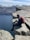 A person with a red backpack sits on the edge of a steep, rocky cliff overlooking a vast, deep-blue body of water. Snow-capped mountains stretch across the background under a bright, clear sky. The scene conveys a sense of adventure and tranquility.