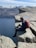 A person with a red backpack sits on the edge of a steep, rocky cliff overlooking a vast, deep-blue body of water. Snow-capped mountains stretch across the background under a bright, clear sky. The scene conveys a sense of adventure and tranquility.
