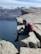 A person with a red backpack sits on the edge of a steep, rocky cliff overlooking a vast, deep-blue body of water. Snow-capped mountains stretch across the background under a bright, clear sky. The scene conveys a sense of adventure and tranquility.