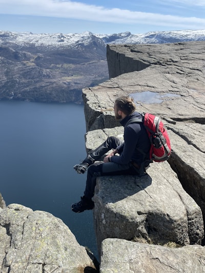 A person with a red backpack sits on the edge of a steep, rocky cliff overlooking a vast, deep-blue body of water. Snow-capped mountains stretch across the background under a bright, clear sky. The scene conveys a sense of adventure and tranquility.