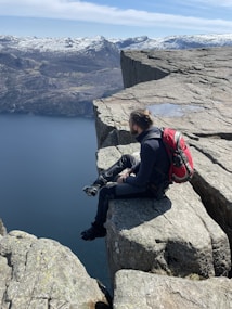 A person with a red backpack sits on the edge of a steep, rocky cliff overlooking a vast, deep-blue body of water. Snow-capped mountains stretch across the background under a bright, clear sky. The scene conveys a sense of adventure and tranquility.