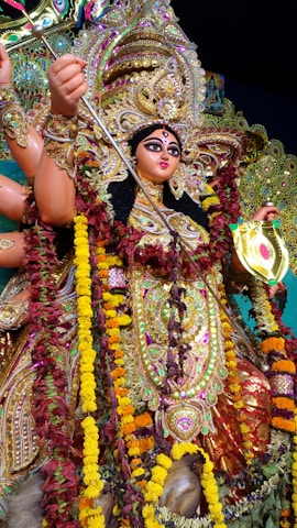 A richly adorned statue of a goddess with multiple arms features elaborate jewelry and vibrant attire. Intricate patterns and decorations are present, including colorful garlands of flowers. The figure holds a weapon and appears in a dynamic and powerful pose.