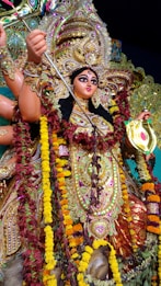 A richly adorned statue of a goddess with multiple arms features elaborate jewelry and vibrant attire. Intricate patterns and decorations are present, including colorful garlands of flowers. The figure holds a weapon and appears in a dynamic and powerful pose.