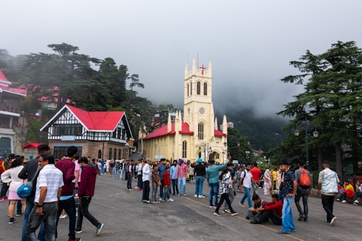 A large crowd of people gathers in front of a historic church with a tall clock tower and red roof. The church is surrounded by lush green trees, and the sky is overcast with a layer of fog gently enveloping the background. Nearby, there are additional buildings with similar architectural styles, featuring exposed brick and red rooftops.