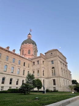 A large, historic government building with a central dome topped by a flag, surrounded by lush greenery and a clear blue sky. The building is made of light-colored stone, and the architectural style includes arched windows and decorative elements. The moon is visible in the sky.