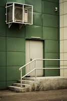 A green-paneled building wall with a closed metal door is featured, accompanied by a set of concrete steps leading to the door. Above the door, an air conditioning unit is secured in a protective cage. The scene conveys an industrial or utilitarian environment.