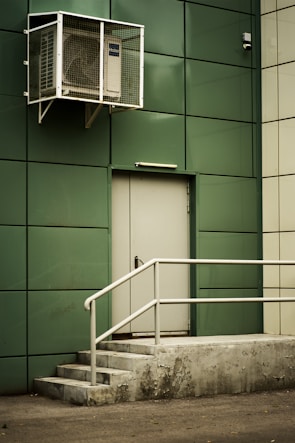 A green-paneled building wall with a closed metal door is featured, accompanied by a set of concrete steps leading to the door. Above the door, an air conditioning unit is secured in a protective cage. The scene conveys an industrial or utilitarian environment.
