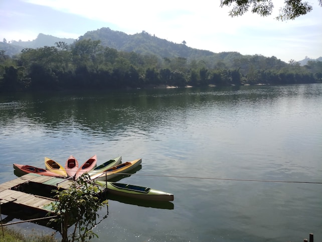 A tranquil scene of a calm river flanked by lush green hills covered in dense trees. Several colorful canoes are neatly lined up at a small wooden dock, with reflections visible in the clear water. The sky is light blue with a few clouds and the overall atmosphere is peaceful and serene.