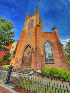 A tall, historic brick church features a prominent arched doorway with ornate wooden double doors. The facade includes large arched windows and is framed by a decorative iron fence. Green bushes and trees provide a natural contrast to the red brick, and the sky is a vibrant blue.