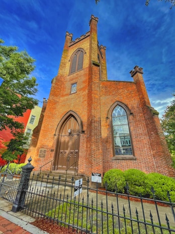A tall, historic brick church features a prominent arched doorway with ornate wooden double doors. The facade includes large arched windows and is framed by a decorative iron fence. Green bushes and trees provide a natural contrast to the red brick, and the sky is a vibrant blue.